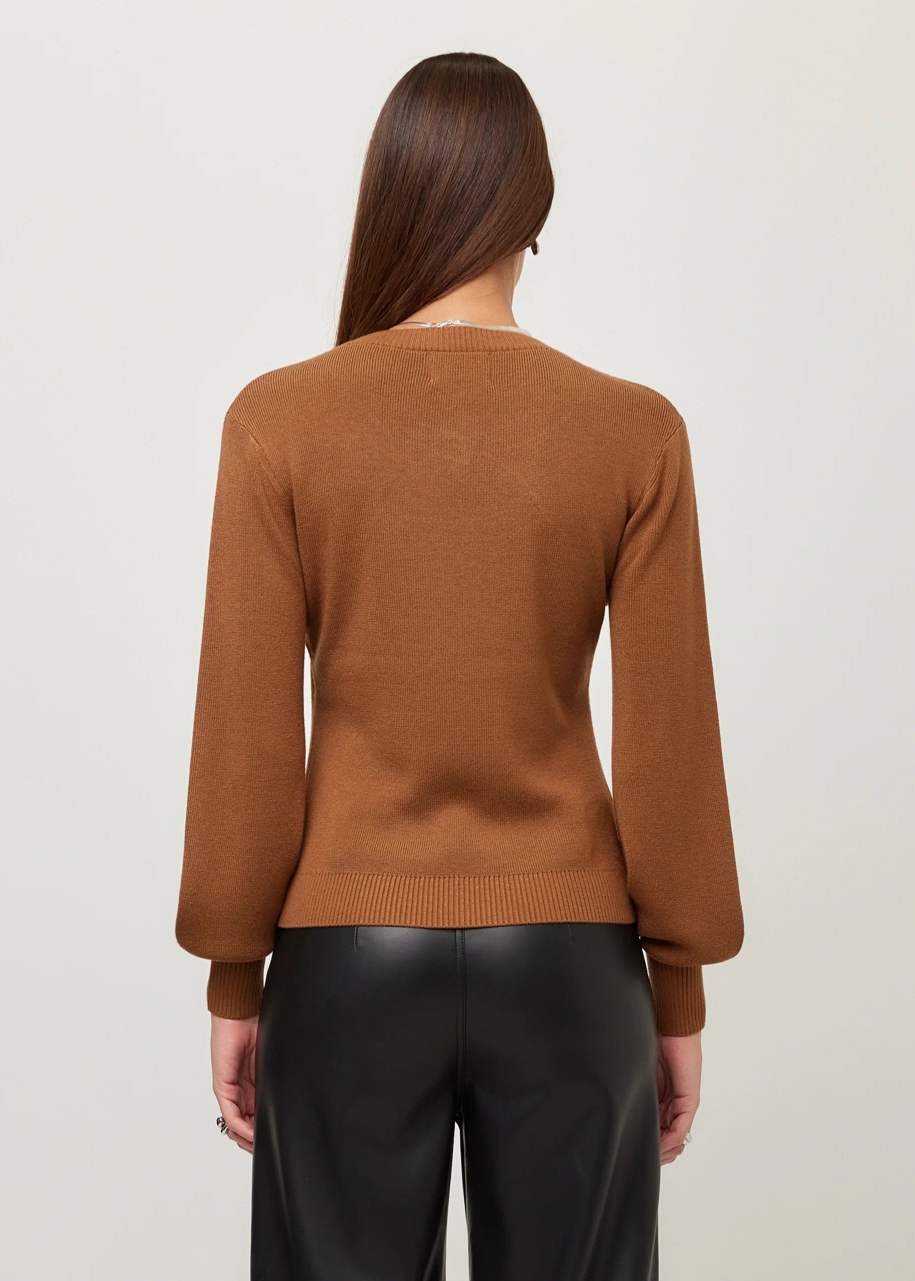 A woman viewed from the back wearing a fitted brown knit sweater and black pants against a plain light gray studio background.