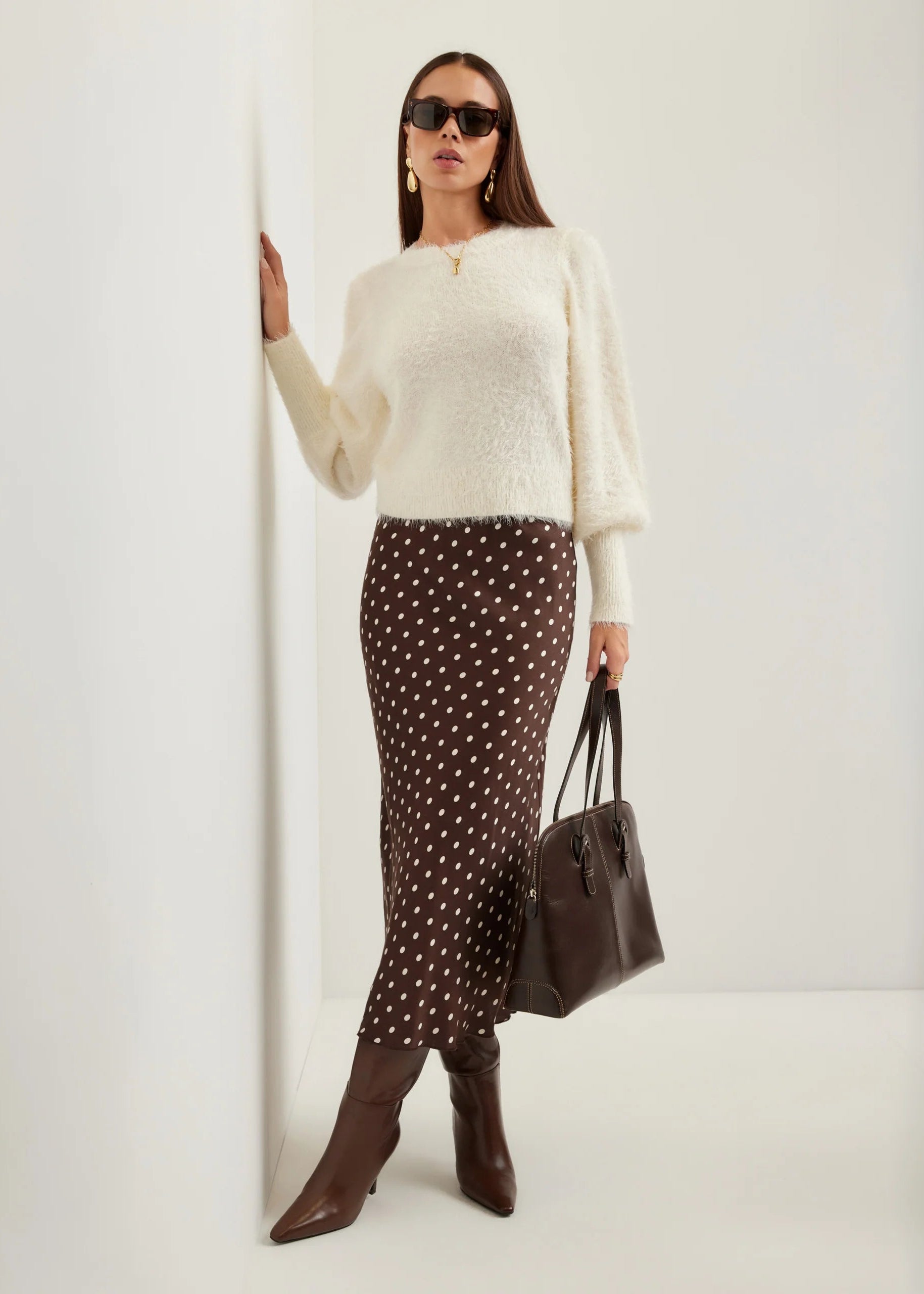 A woman modeling a cream fuzzy long-sleeve sweater, a brown midi skirt with white polka dots, brown pointed-toe ankle boots, and carrying a dark brown leather tote bag against a clean white wall.