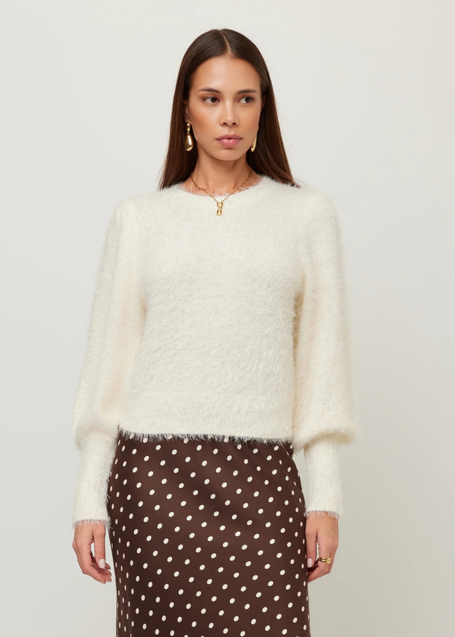 A woman modeling a fuzzy off-white long-sleeve sweater paired with a brown and white polka dot skirt, set against a plain light gray studio background.