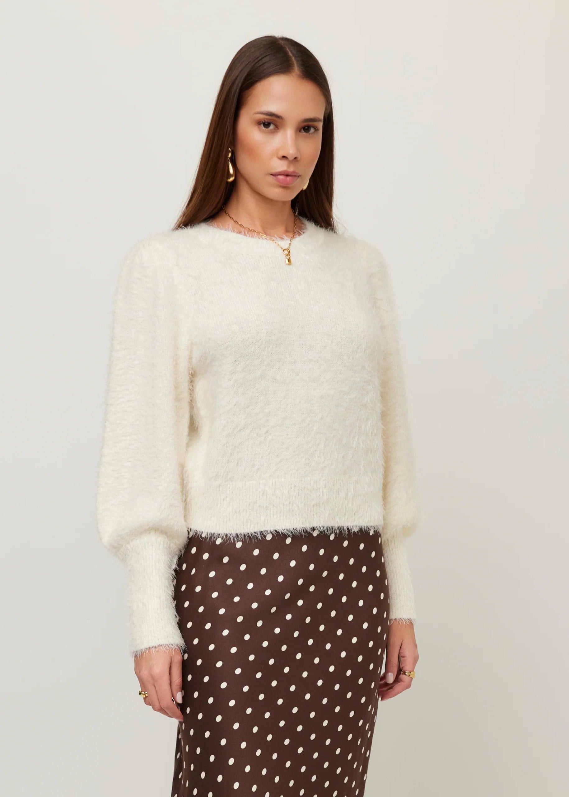 A woman modeling a fuzzy off-white long-sleeve sweater paired with a brown skirt with white polka dots, against a plain light gray background.