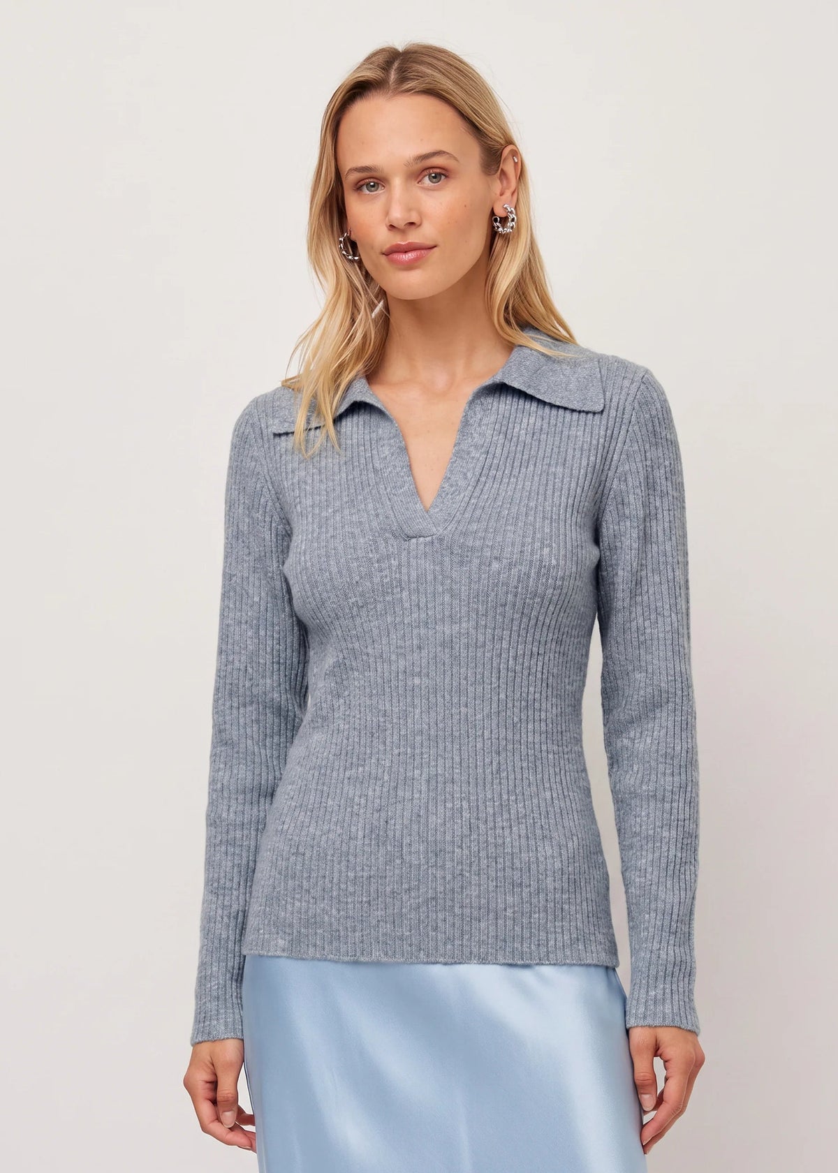 A woman modeling a light gray ribbed knit polo-neck sweater with a split V-neckline, paired with a light blue satin skirt, against a plain off-white studio background.