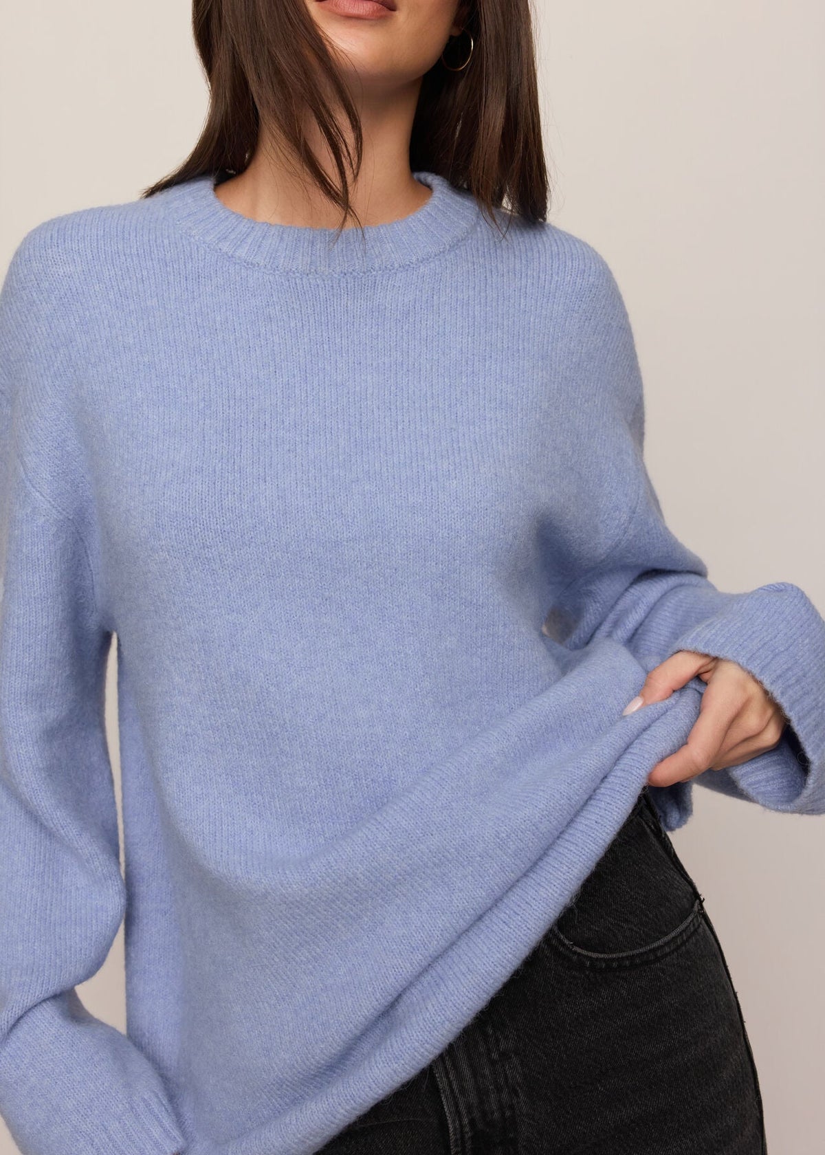 A woman wearing a light blue ribbed knit crewneck sweater and black jeans, shown from the neck down against a plain beige studio background.