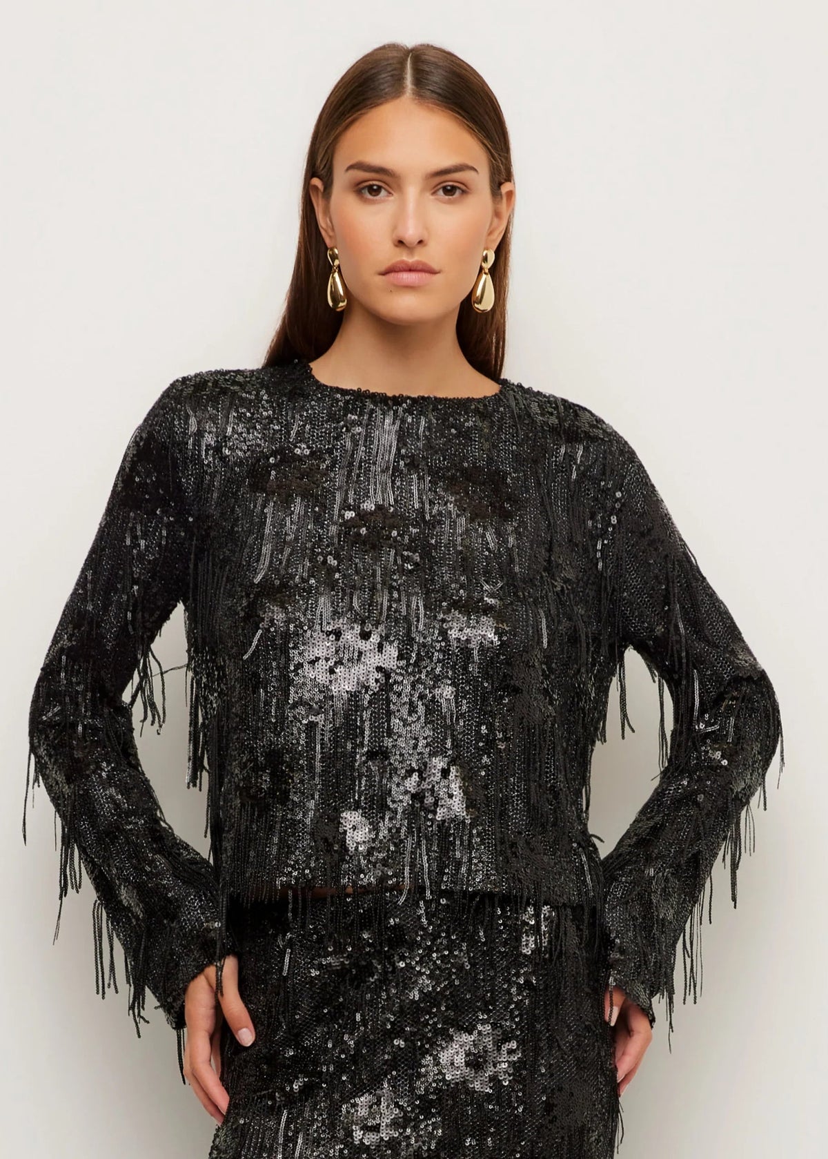 A woman modeling a black long-sleeve top covered in shimmering sequins and fringe, paired with a matching fringed skirt, against a plain light gray studio background.