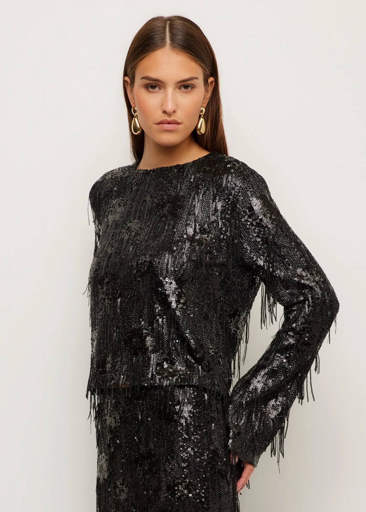 A woman modeling a black long-sleeve sequined dress with fringe detailing, set against a plain light gray studio background.
