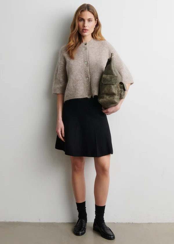 Woman wearing a beige sweater, black skirt, and black shoes holding a green bag against a white background