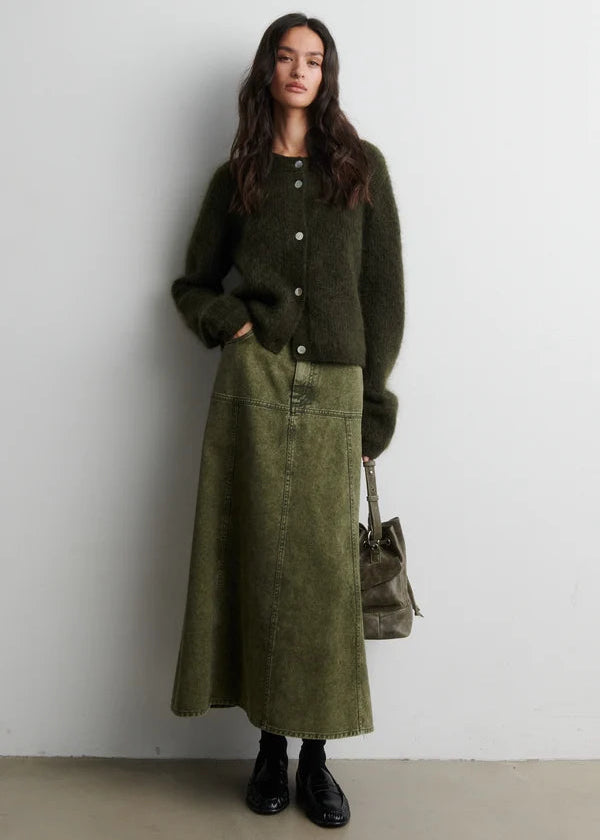Woman wearing a green outfit with a long coat and skirt, holding a bag against a white background