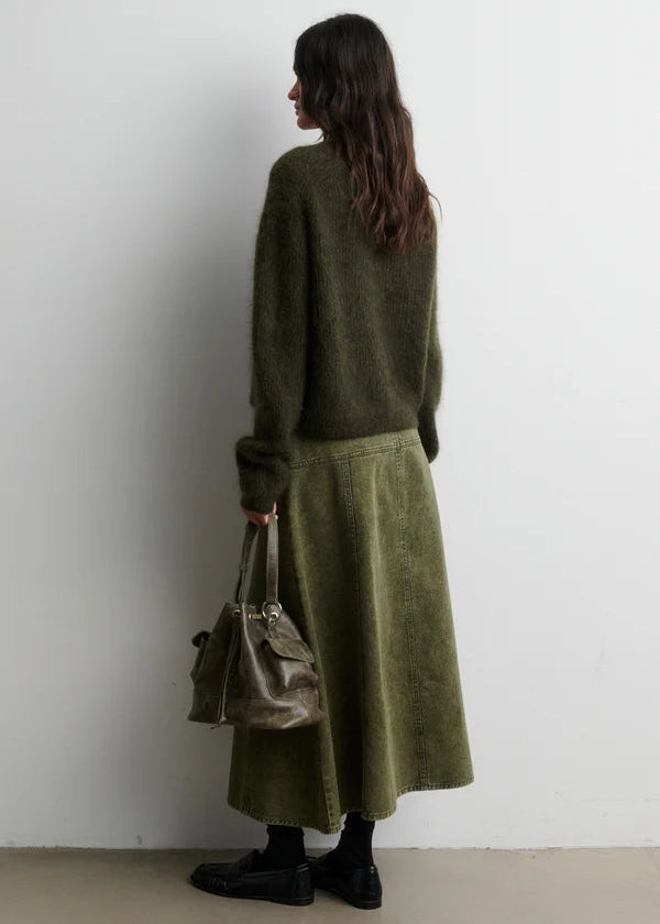 Woman wearing a green outfit with a long skirt and sweater, holding a beige handbag against a white wall.