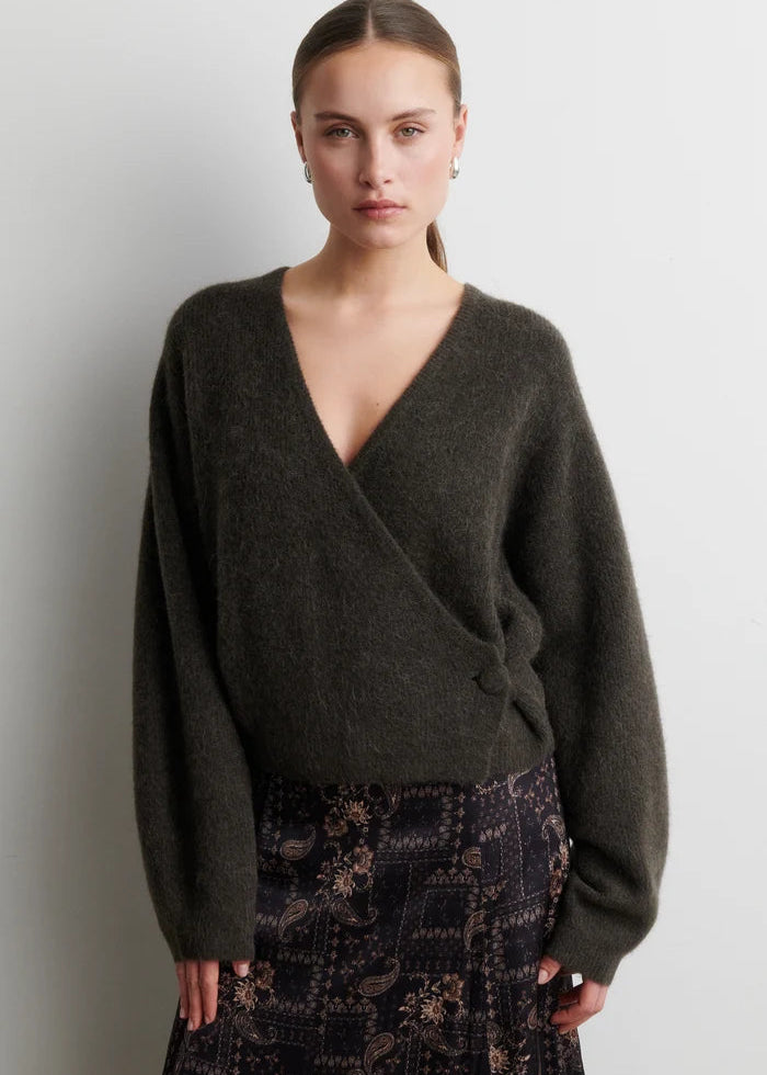 A woman modeling a dark olive green wrap-style knit sweater with a deep V-neck and long sleeves, paired with a dark patterned skirt, against a plain light gray wall.