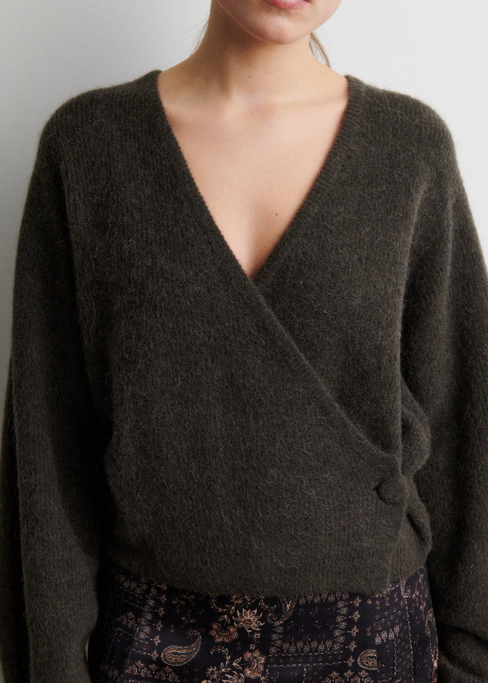 A close-up of a person wearing a dark olive green fuzzy knit wrap sweater with a deep V-neck and side button detail, paired with patterned pants.