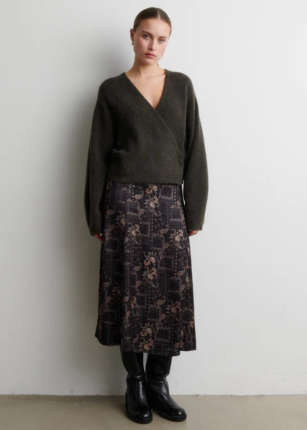 A model stands against a plain light gray wall wearing a dark olive wrap sweater, a dark patterned midi skirt, and black boots.