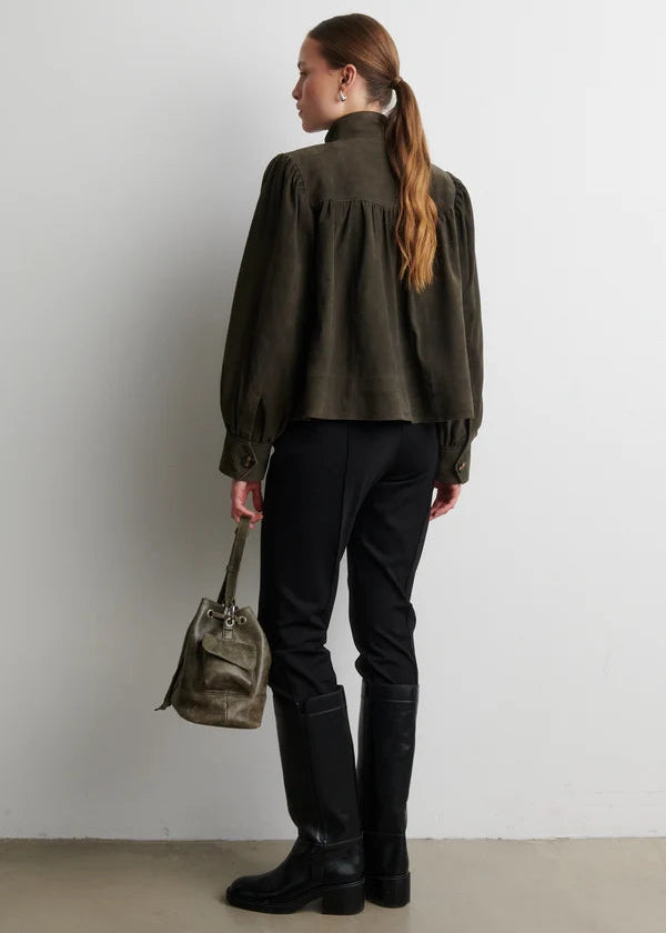 A woman stands with her back to the camera wearing an olive green long-sleeve blouse with gathered shoulders, black pants, and black knee-high boots, holding a matching olive green handbag against a plain light gray wall.