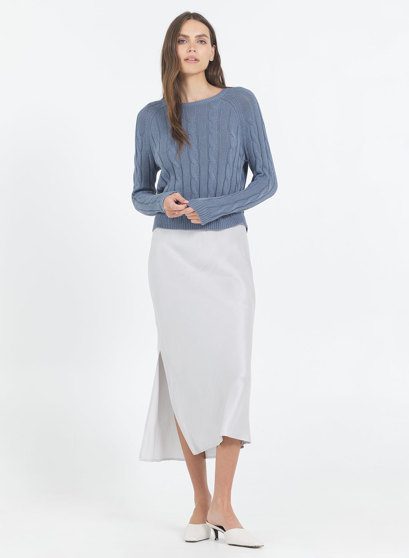 Woman wearing the One Grey Day blue Raleigh Cable sweater in Cadet and white skirt on a white background.
