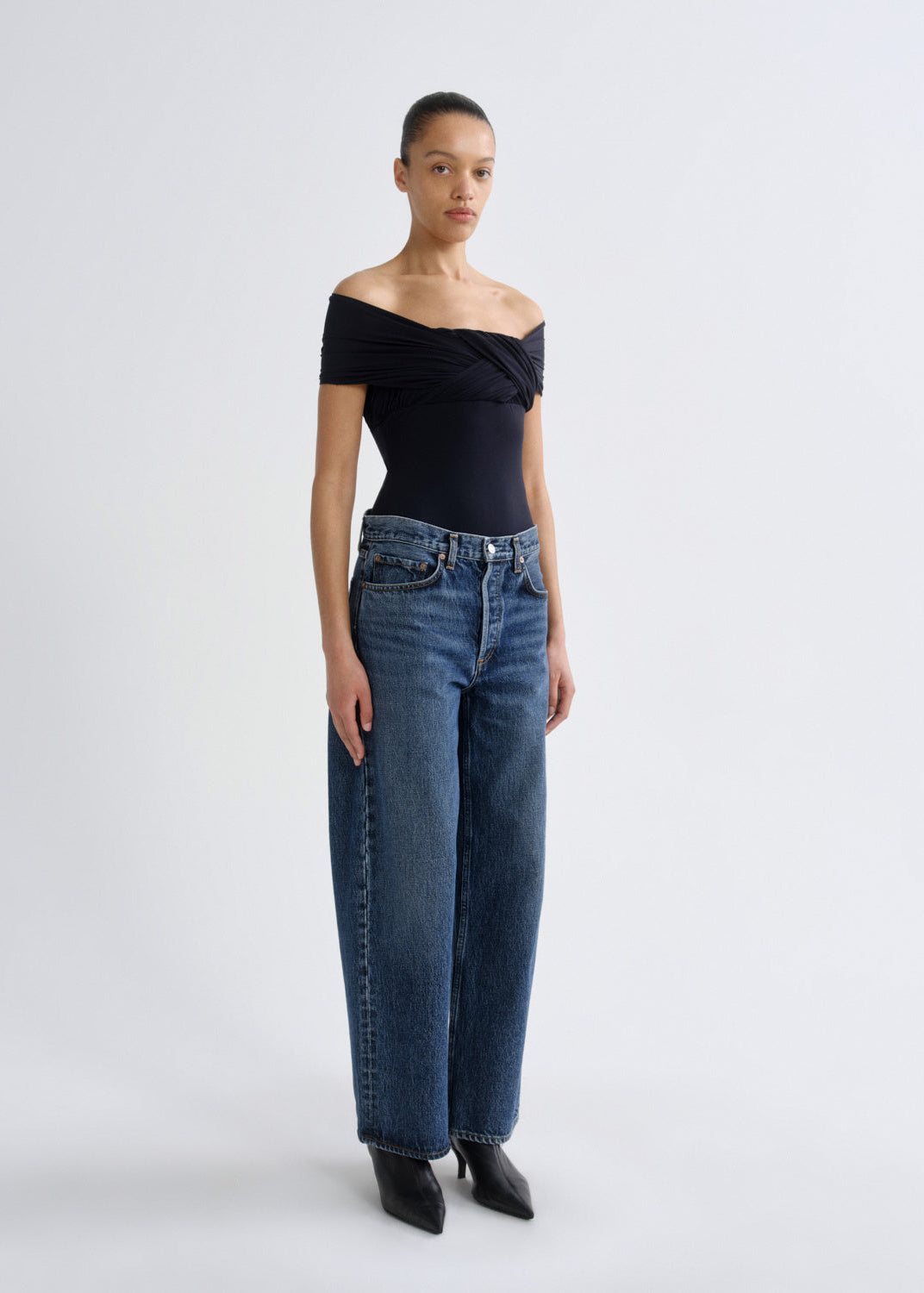 A model stands against a light gray studio background wearing a black off-the-shoulder top, blue wide-leg jeans, and black pointed shoes.