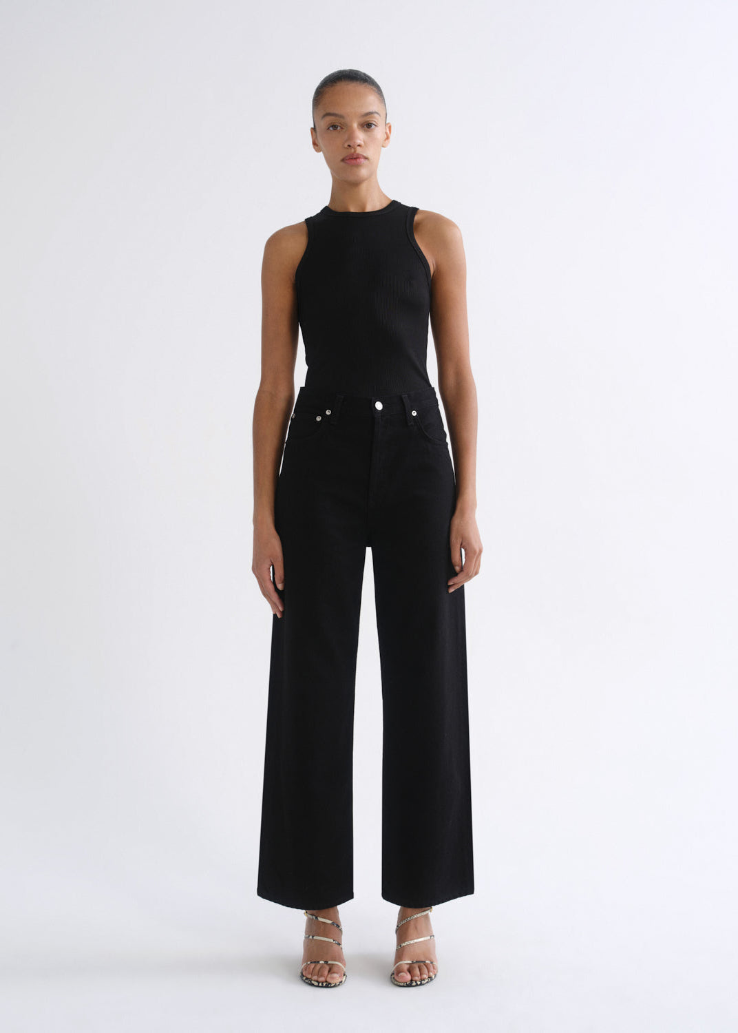 A model stands against a plain white studio background wearing a black sleeveless high-neck top and matching black wide-leg pants with silver-tone hardware, paired with beige heeled sandals.
