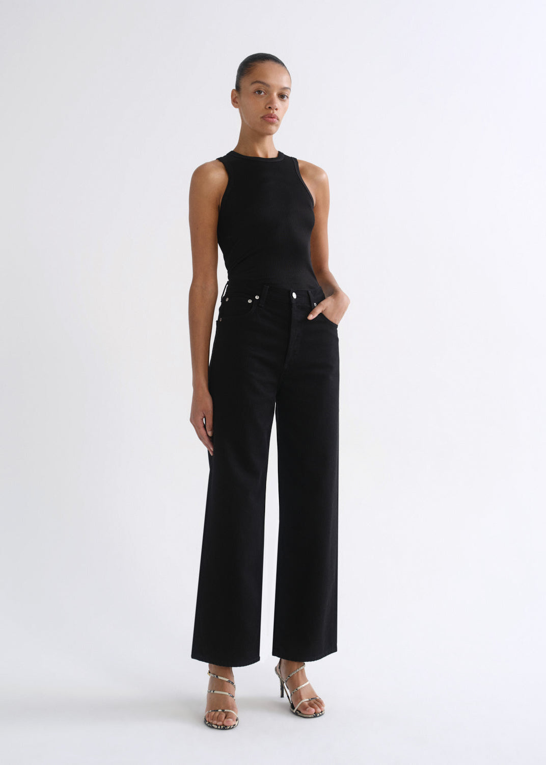 A model stands against a plain white studio background wearing a black sleeveless top and black wide-leg cropped pants, paired with metallic heeled sandals.