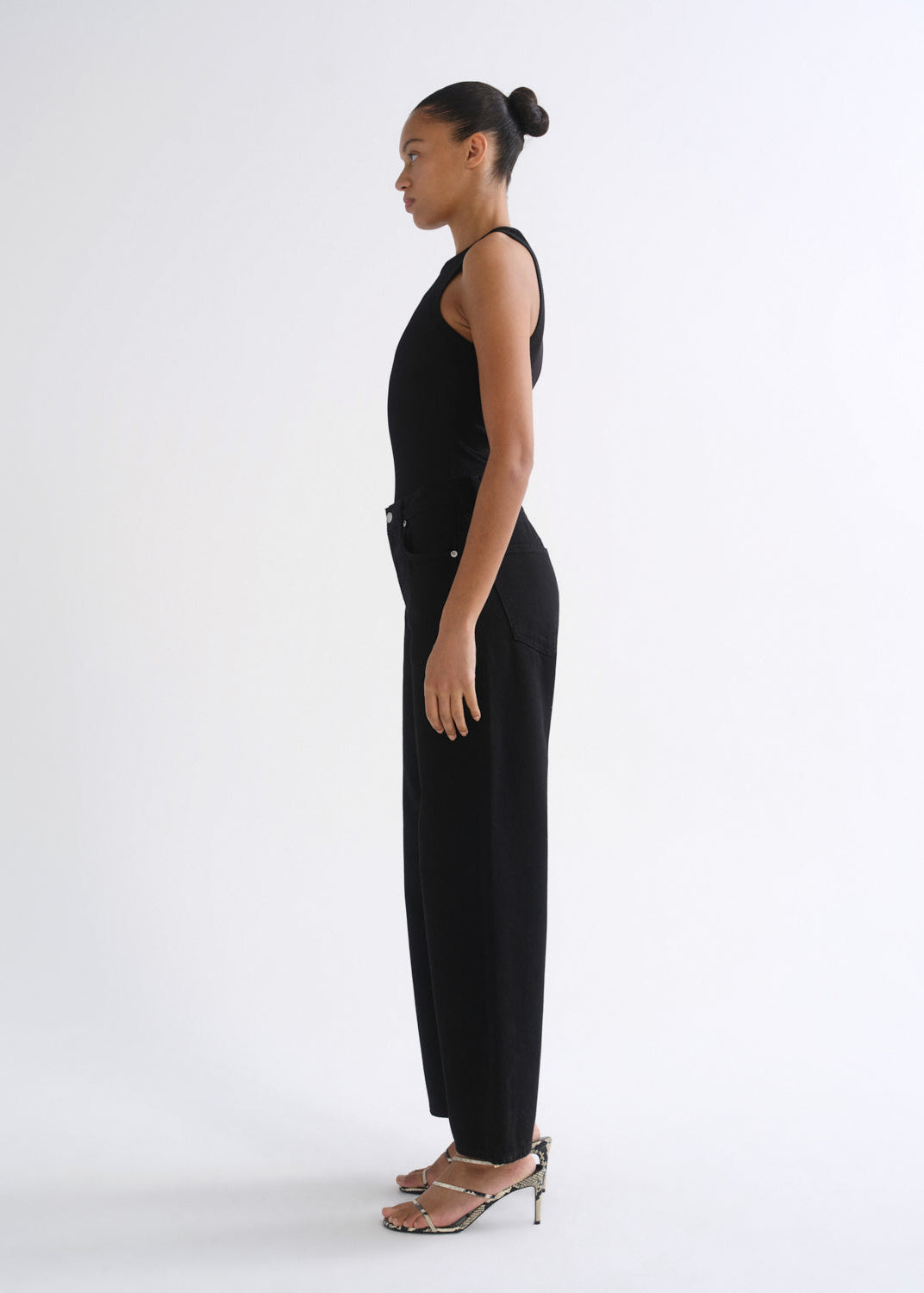 A woman in a black sleeveless jumpsuit and metallic heeled sandals, shown in profile against a plain white studio background.