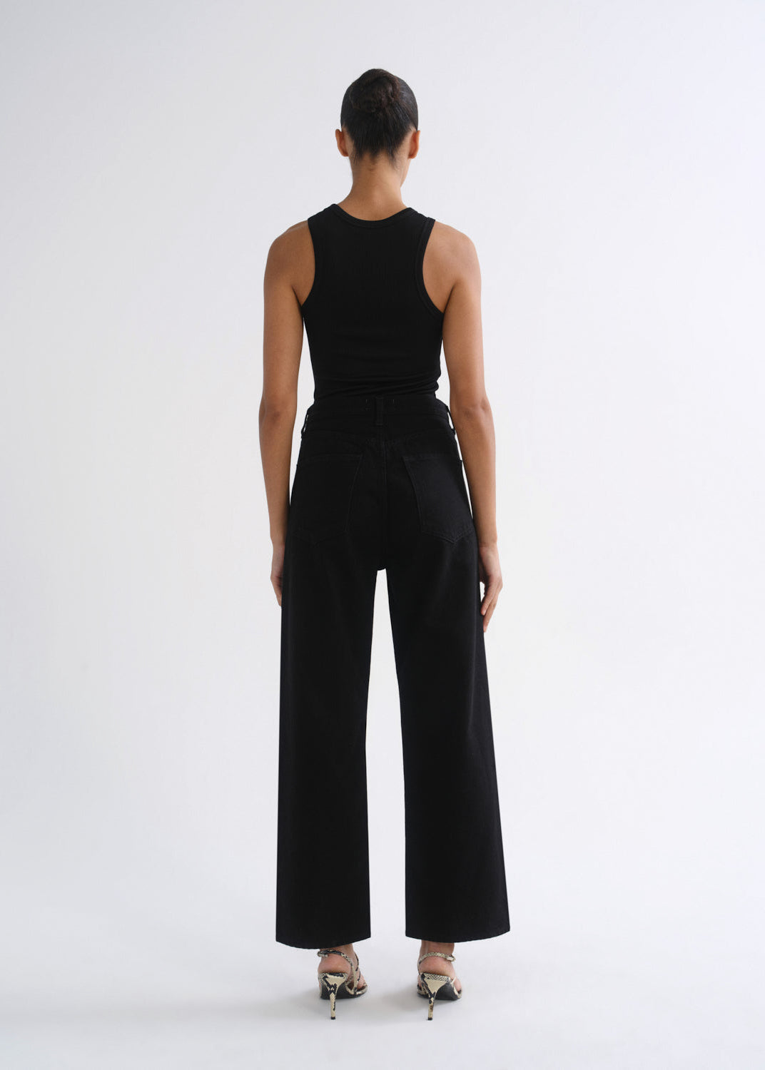 A model viewed from the back wearing a black sleeveless top and black wide-leg trousers, paired with metallic high-heeled sandals, against a plain white studio background.