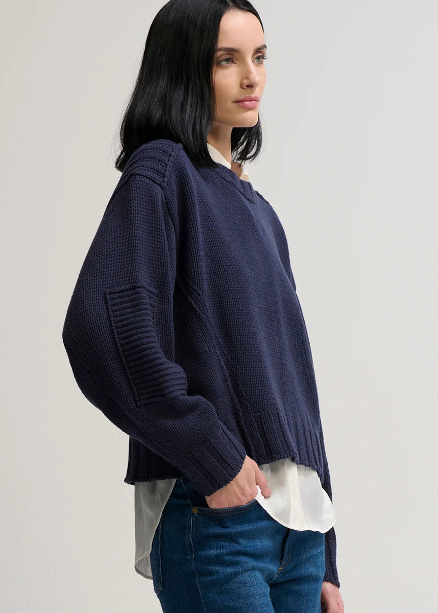 A woman wearing a dark navy blue knit sweater with ribbed details on the sleeves, layered over a white collared shirt and paired with blue jeans, photographed against a neutral light gray background.
