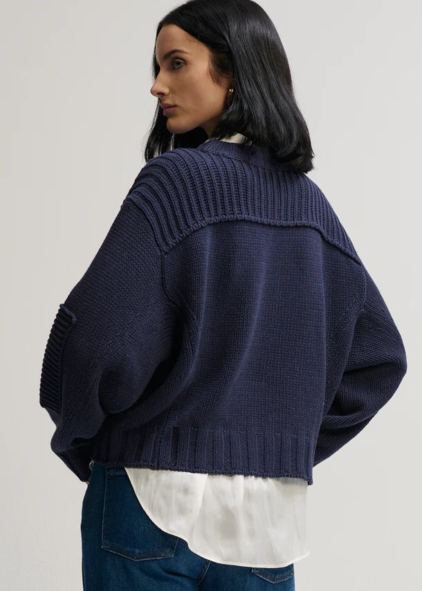A woman shown from the back wearing a cropped navy blue knit cardigan over a white shirt and blue jeans, photographed against a plain light gray studio background.