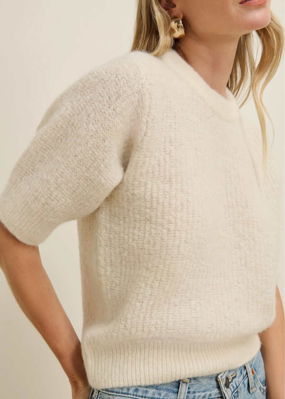 A close-up of a woman wearing a short-sleeve knit sweater in a warm off-white or cream color, paired with blue jeans and gold earrings, against a plain light beige background.