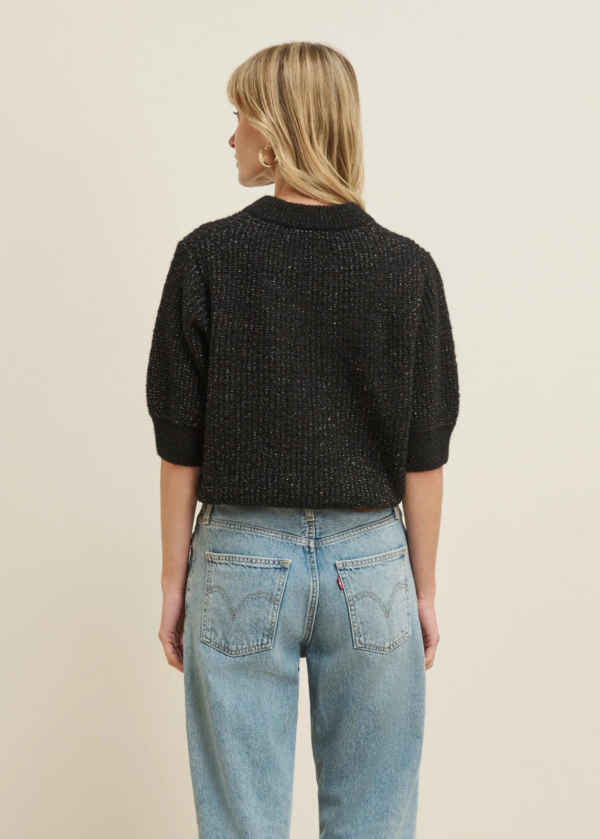 A woman viewed from the back wearing a black textured knit sweater and light blue jeans, standing against a plain beige studio background.
