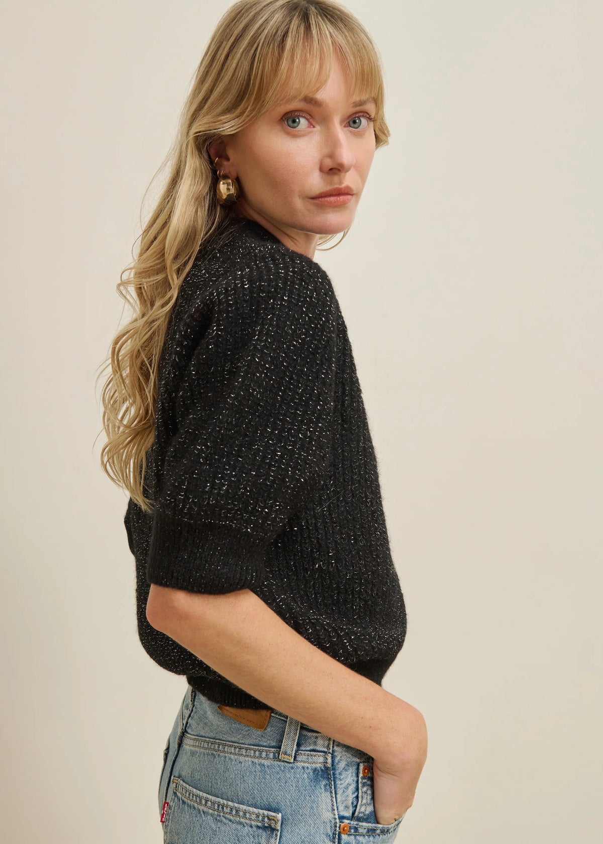 A woman with blonde hair wearing a black textured knit sweater and light blue jeans, photographed from the side against a plain beige background.