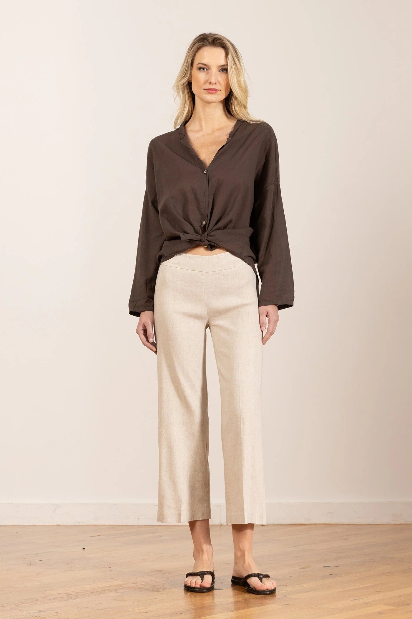 Woman wearing a brown blouse and beige pants standing against a plain background