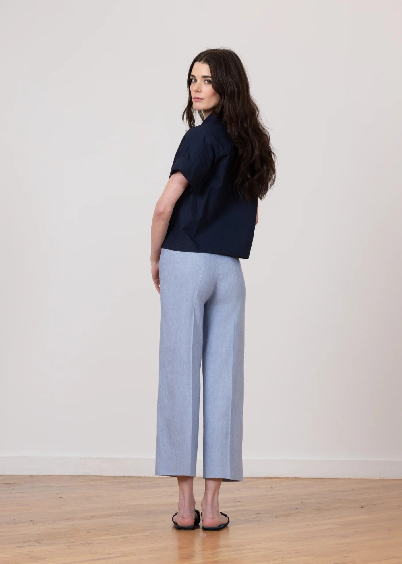 Woman wearing a navy shirt and light gray pants standing on a wooden floor with a white wall background
