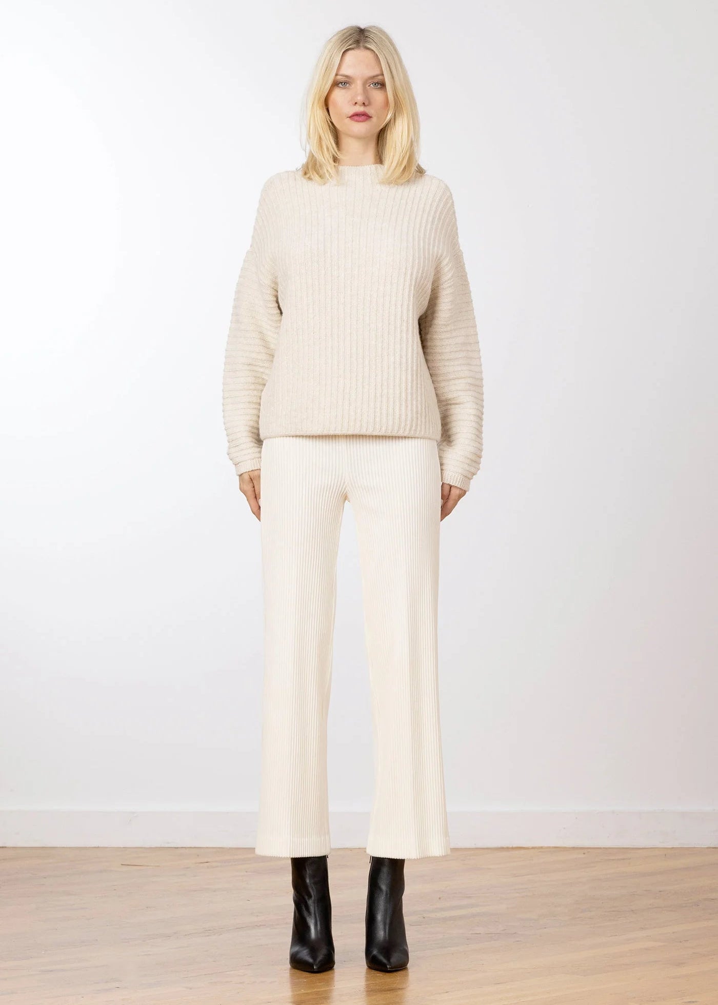 A blonde model stands in a studio wearing a cream ribbed knit mock-neck sweater and matching cream wide-leg cropped trousers, paired with black ankle boots.