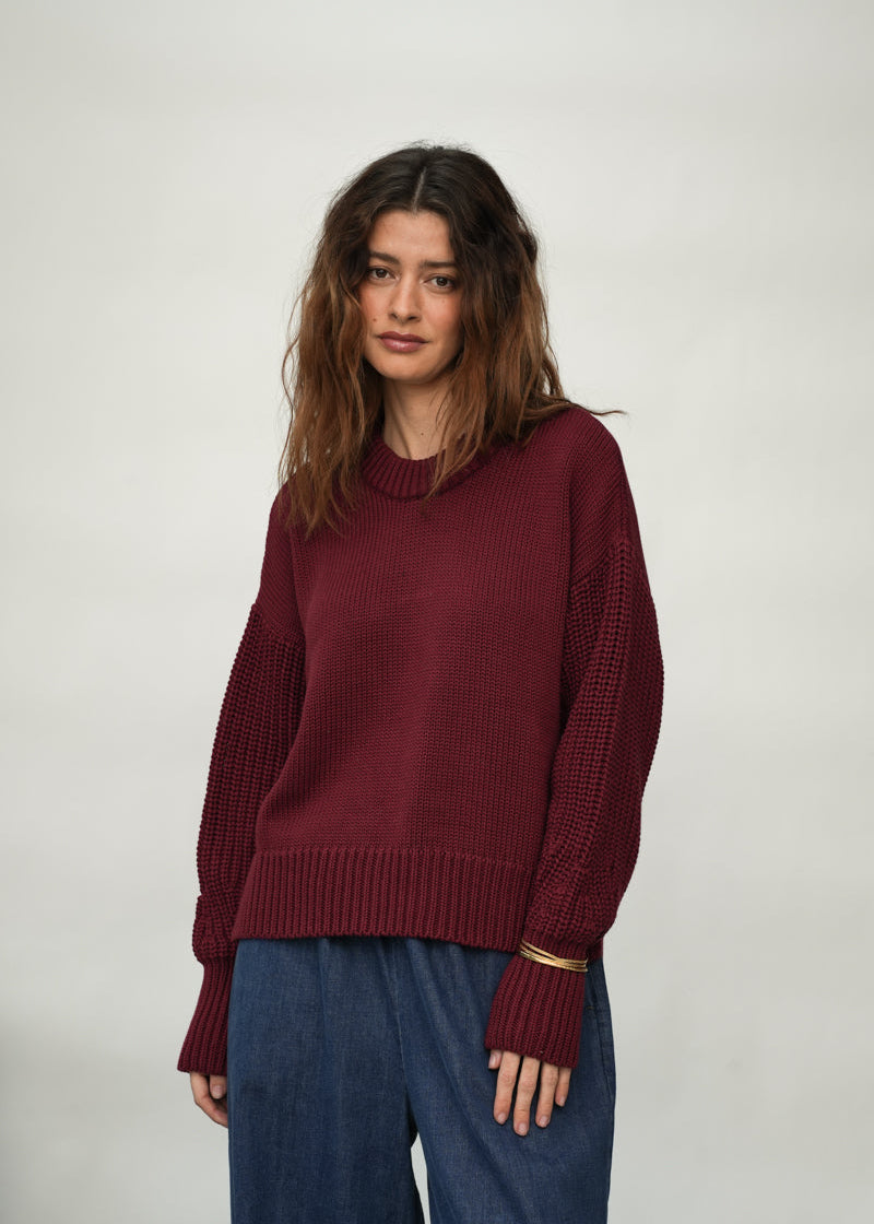 A woman wearing a burgundy knit sweater with textured, ribbed sleeves and a ribbed hem, paired with blue jeans, photographed against a neutral light gray studio background.