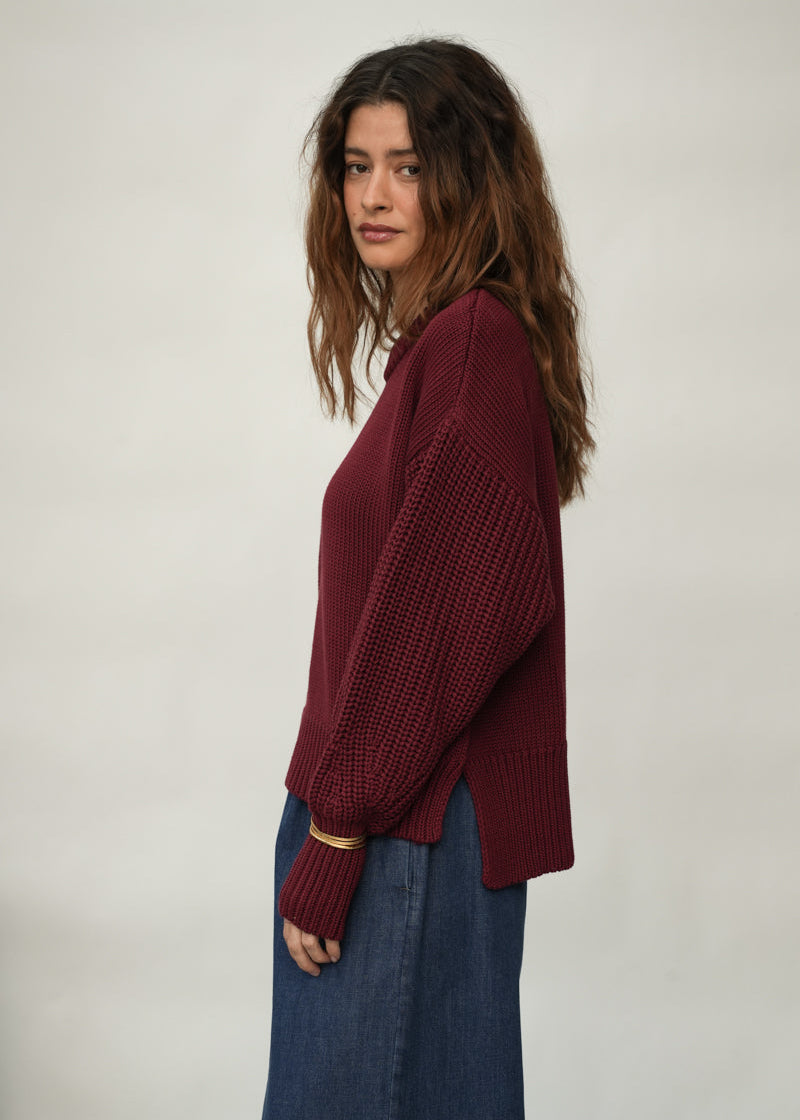 A woman wearing a burgundy knit sweater with textured, ribbed sleeves and a relaxed fit, paired with a long blue skirt, photographed against a plain light gray studio background.
