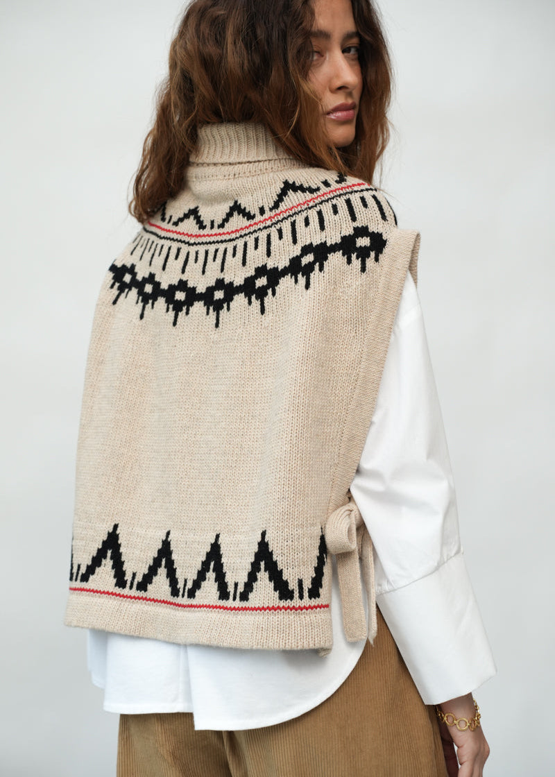 A woman wearing a beige knit turtleneck vest with black geometric patterns and thin red stripes, layered over a white long-sleeve shirt and brown pants, shown from the back against a light gray studio background.