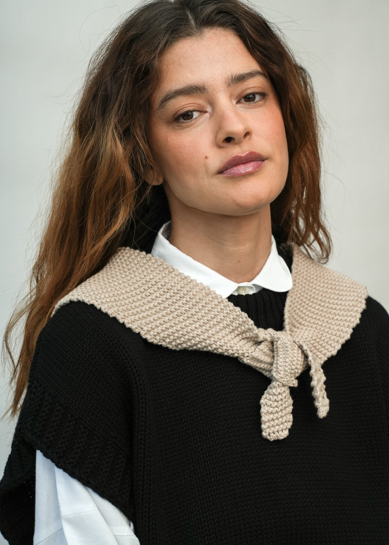 A close-up portrait of a woman wearing a black knit sweater with a wide beige knitted collar tied at the front, layered over a white collared shirt, against a plain light gray background.