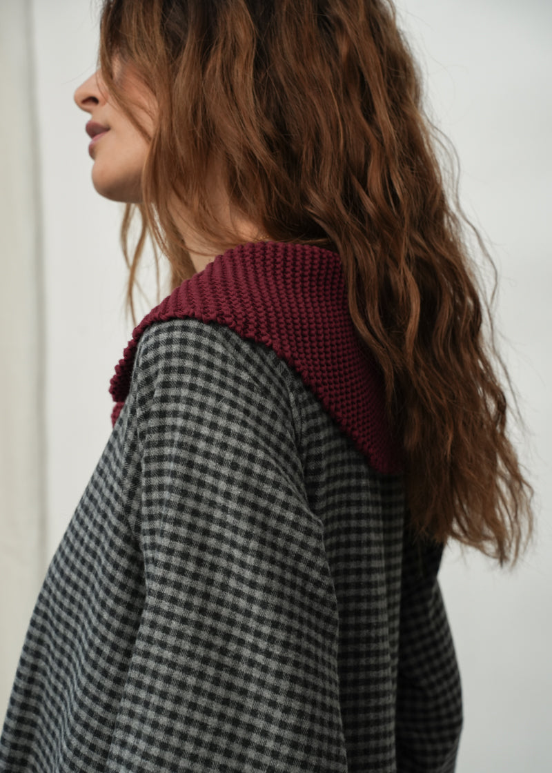 A woman shown in profile wearing a dark gray and black checkered jacket with a contrasting deep burgundy knit collar, set against a plain light background.