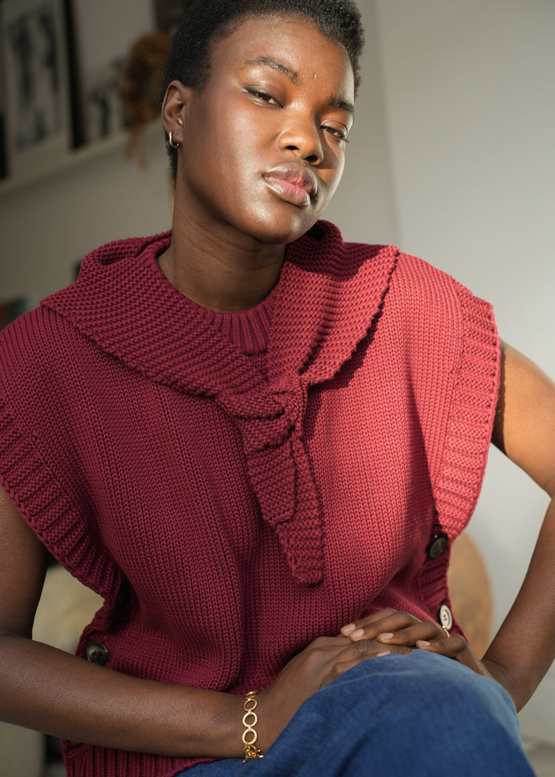 A woman wearing a sleeveless, ribbed knit top in a deep burgundy red, paired with blue jeans. The top features a large ruffled collar tied in a bow at the chest.