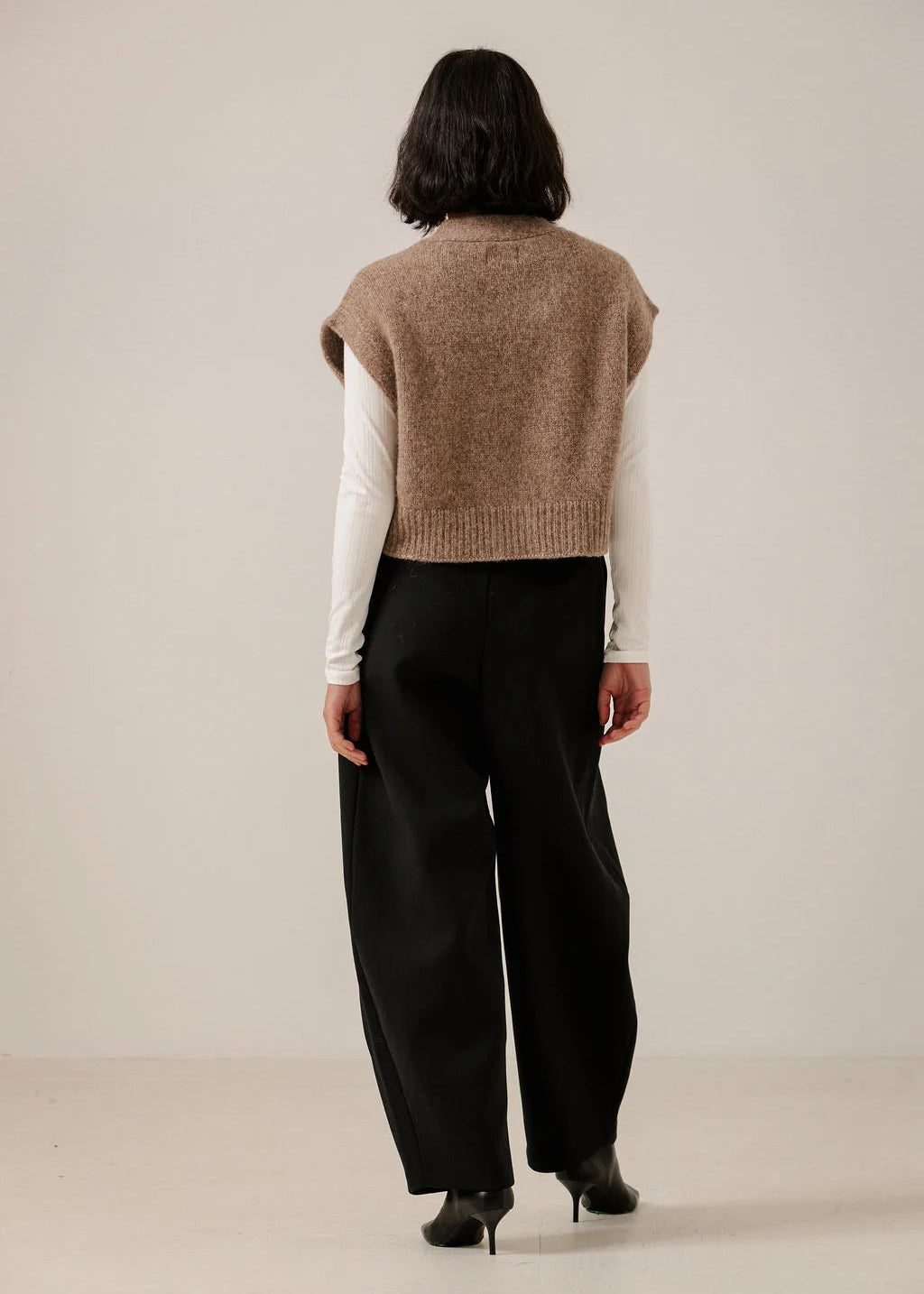 A model shown from the back wearing a sleeveless taupe knit sweater vest over a white long-sleeve shirt and black wide-leg trousers, standing against a plain light beige studio background.