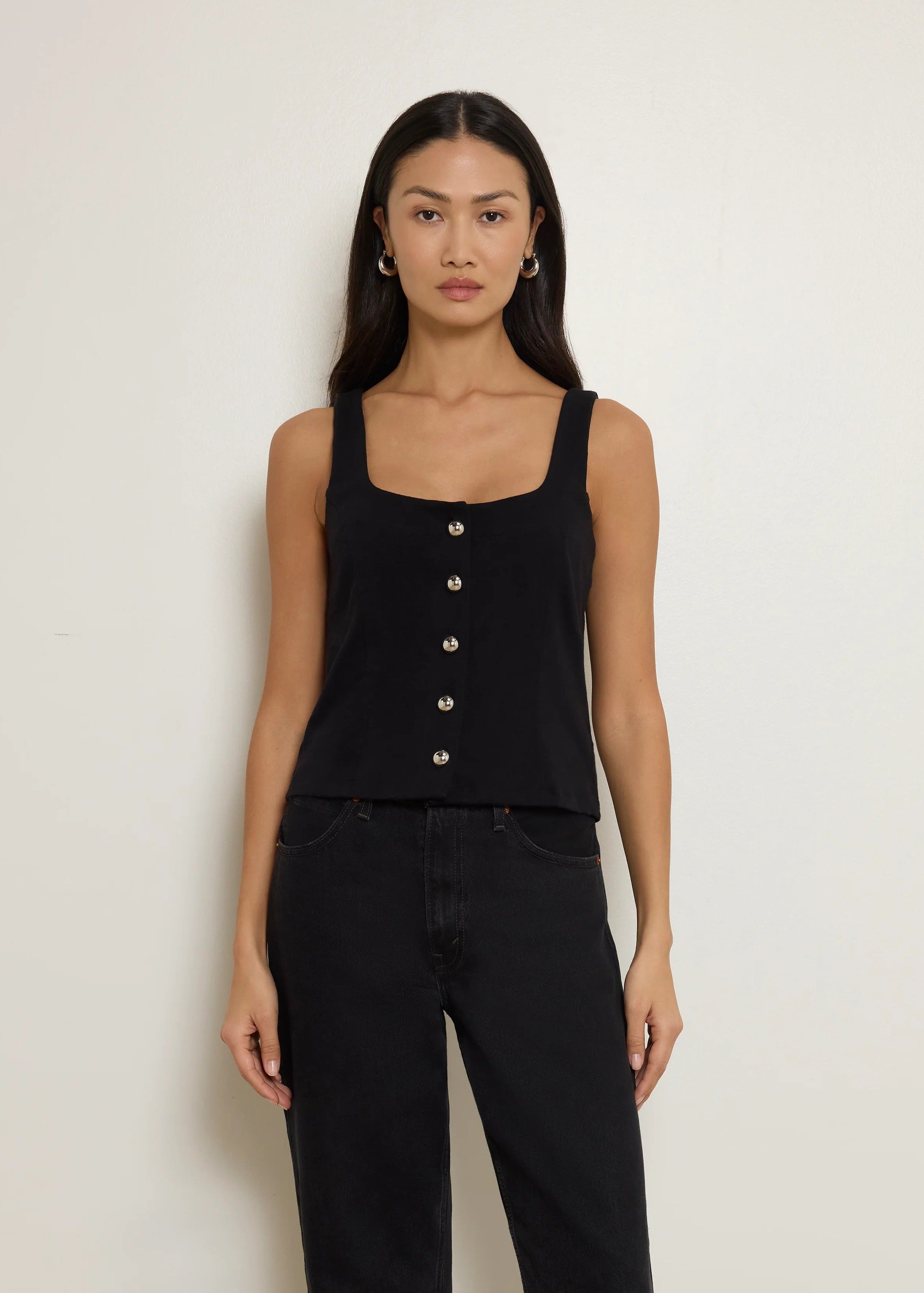 Woman wearing a black sleeveless top with buttons and black pants against a plain background