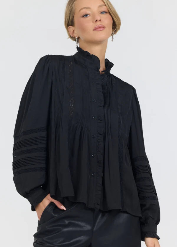 A woman wearing a black long-sleeve blouse with a high ruffled collar, button front, and gathered sleeves, paired with black pants, against a light gray studio background.