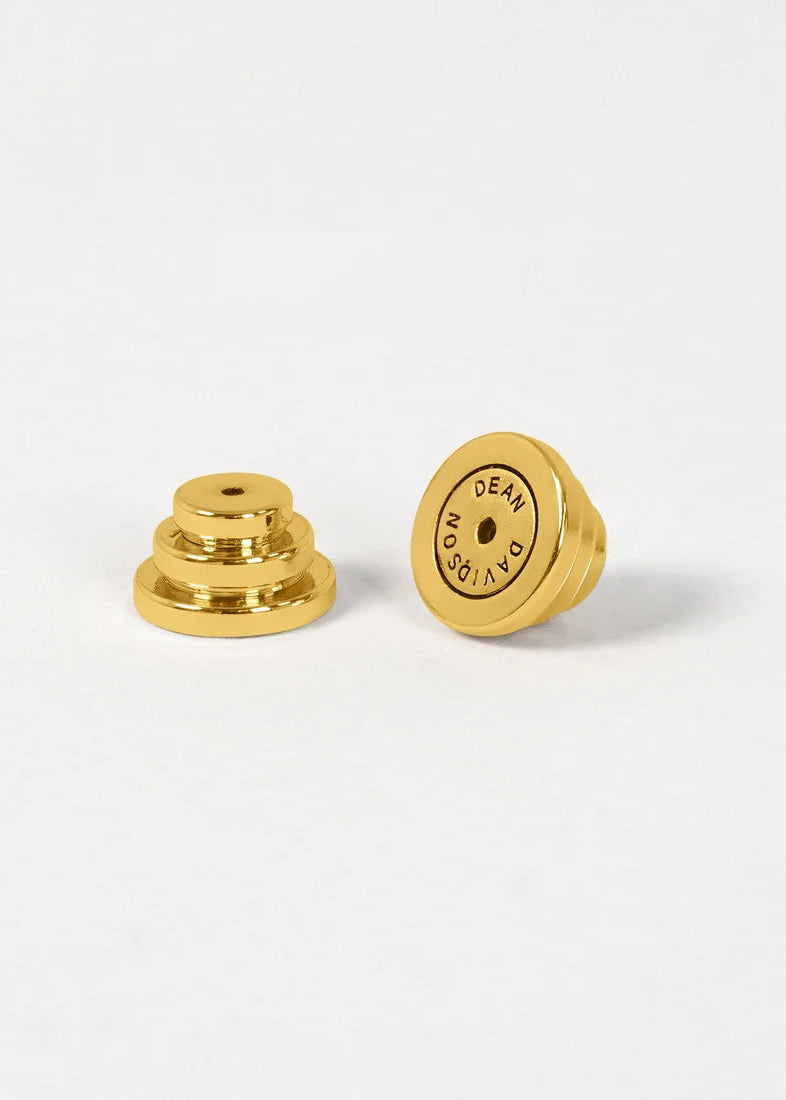 Two small gold metal earbud tips, one showing the layered stem side and the other showing the flat circular end with 'DEAD AIR' printed in black, on a clean white background.