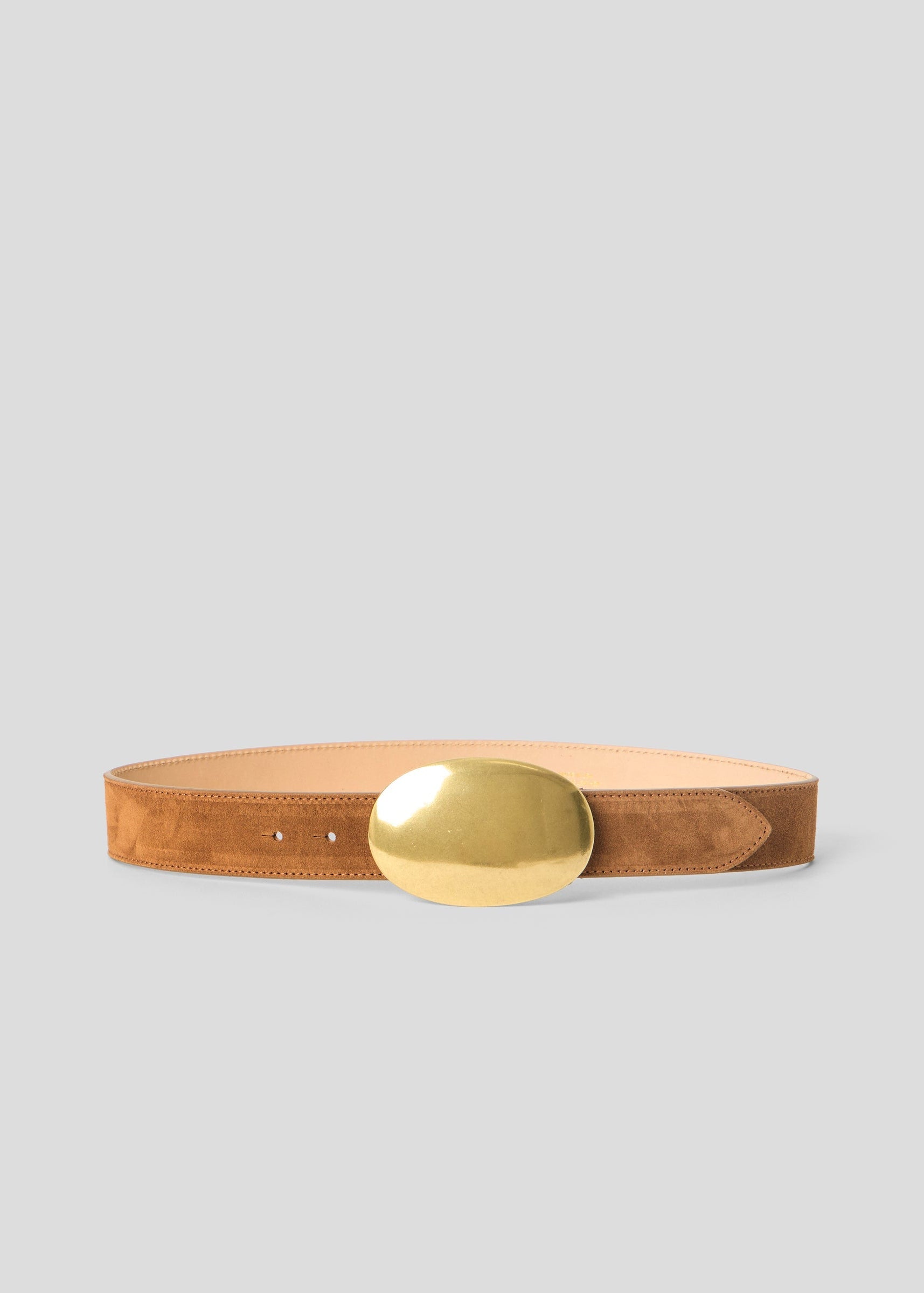 A tan suede belt with a large, polished gold oval buckle, displayed horizontally on a neutral light gray background.