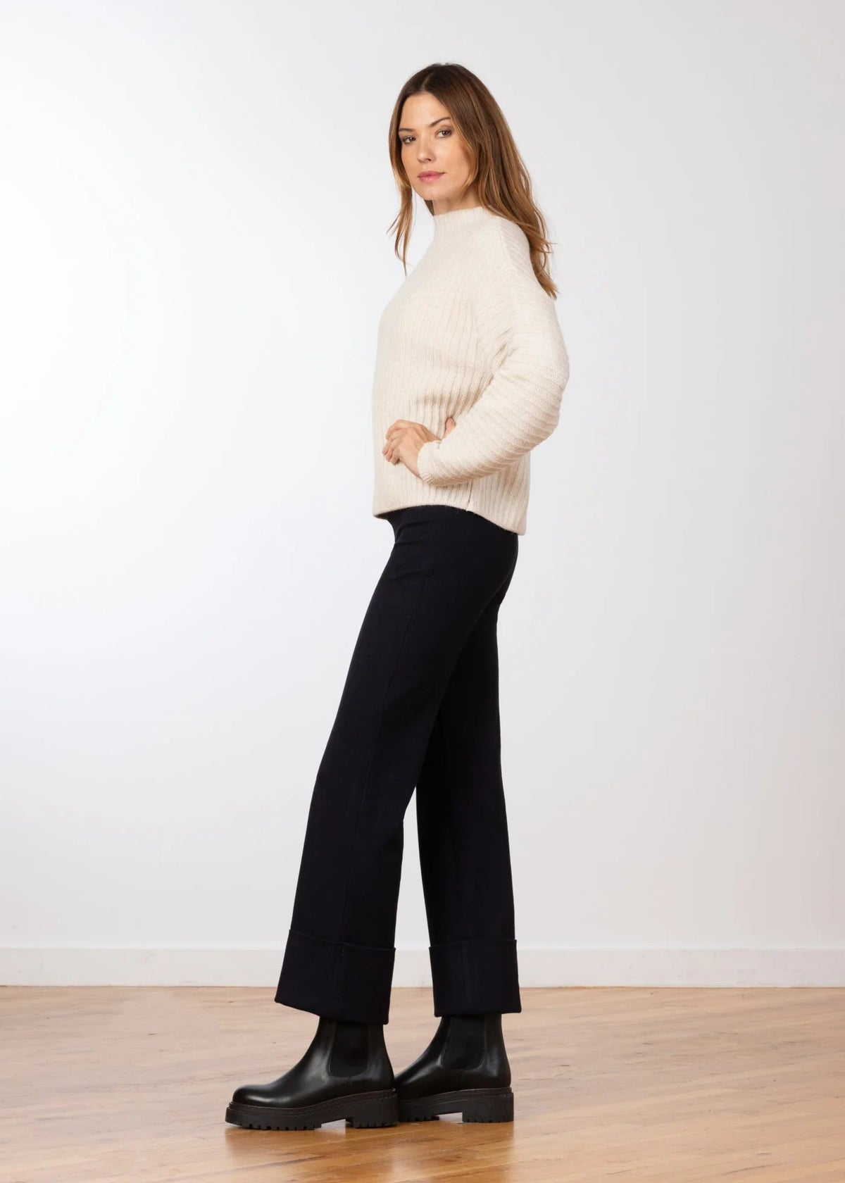A woman modeling a cream ribbed knit mock-neck sweater and black wide-leg cropped pants with black leather boots, standing on a light wood floor against a white wall.