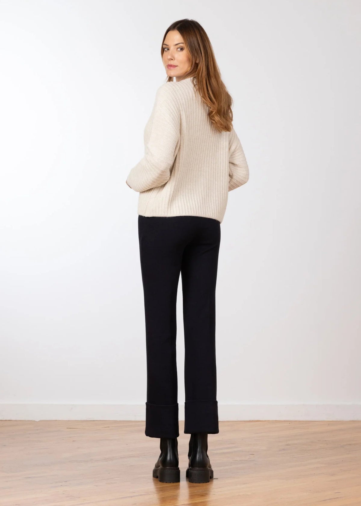 A woman stands with her back to the camera wearing a cream ribbed knit mock-neck sweater, black cropped trousers, and black boots, set against a plain white wall and light wood floor.