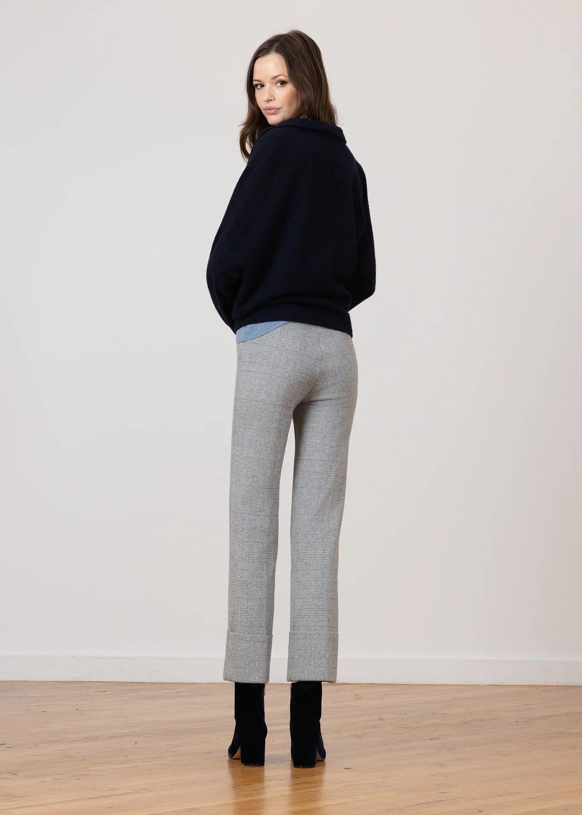 A woman stands with her back to the camera wearing a dark navy sweater, light gray cropped trousers, and black ankle boots in a minimalist room with a light wall and wooden floor.