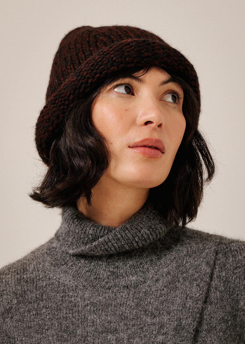 A woman wearing a dark brown chunky knit slouchy beanie and a gray turtleneck sweater, looking off to the side against a plain light beige background.