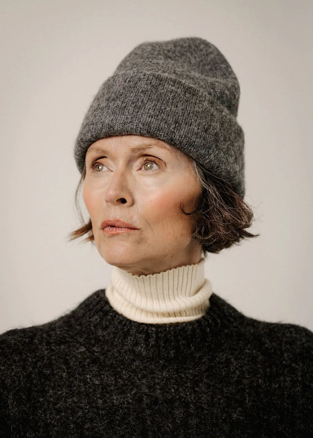 A close-up portrait of a woman wearing a dark gray knit beanie, a cream ribbed turtleneck sweater, and a dark textured sweater, set against a plain light gray background.