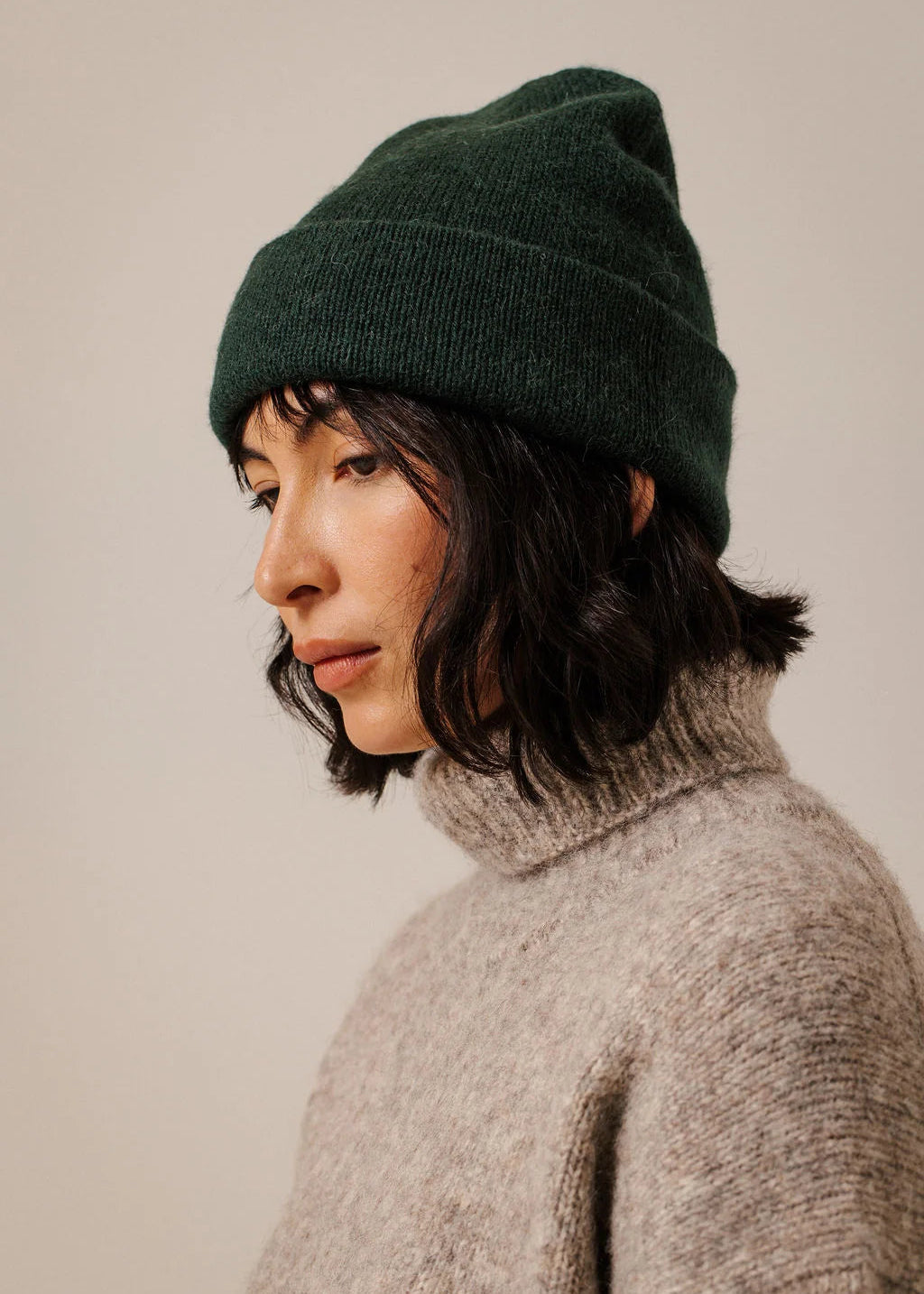 A person shown in profile wearing a dark forest green knit beanie and a light gray turtleneck sweater against a plain beige studio background.