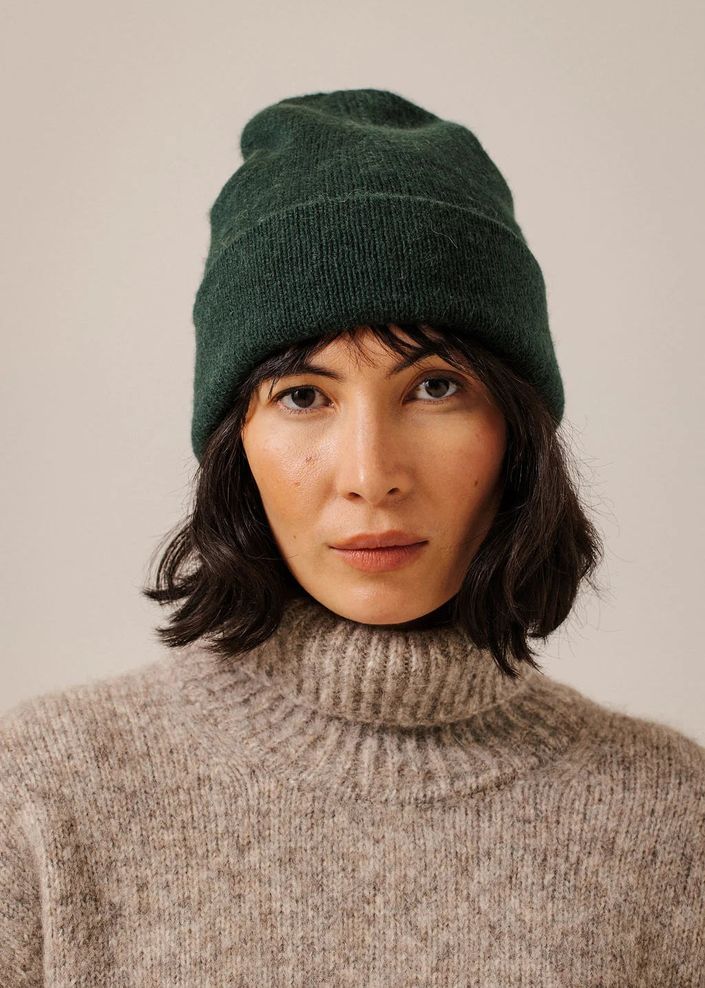 A woman wearing a dark forest green knit beanie and a beige turtleneck sweater, photographed against a plain light beige background.
