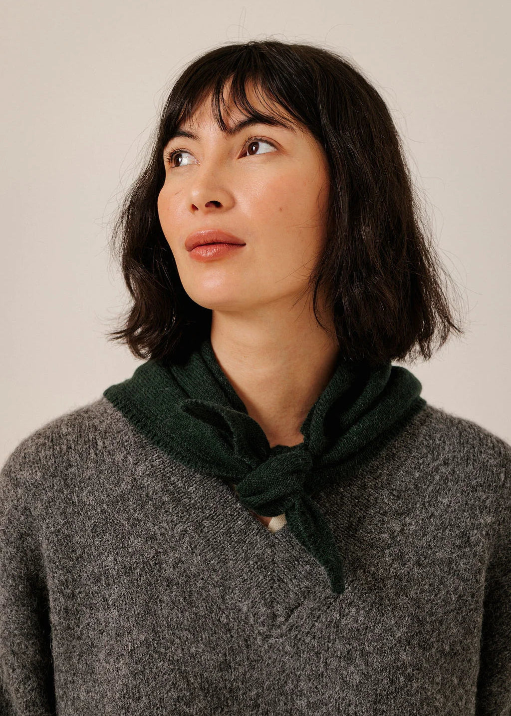 A woman with short dark hair wearing a dark green knitted neck scarf tied at the collar over a gray knit sweater, photographed against a plain light beige background.