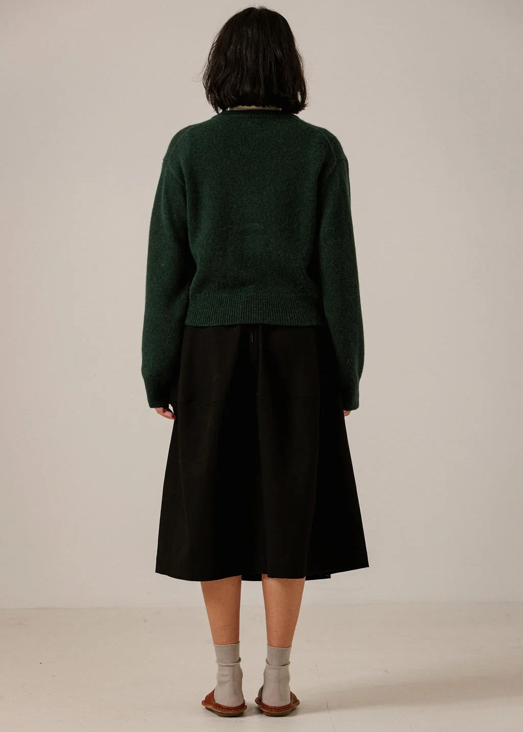 A model viewed from behind wearing a dark green long-sleeve knit sweater and a black midi skirt, paired with light gray socks and brown sandals, against a plain off-white studio background.
