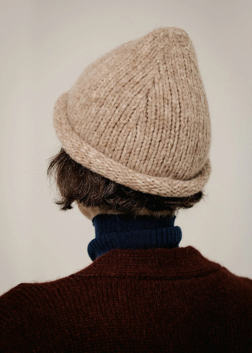 A person shown from behind wearing a beige ribbed knit beanie, a dark navy turtleneck sweater, and a dark brown knit sweater, set against a plain light beige background.